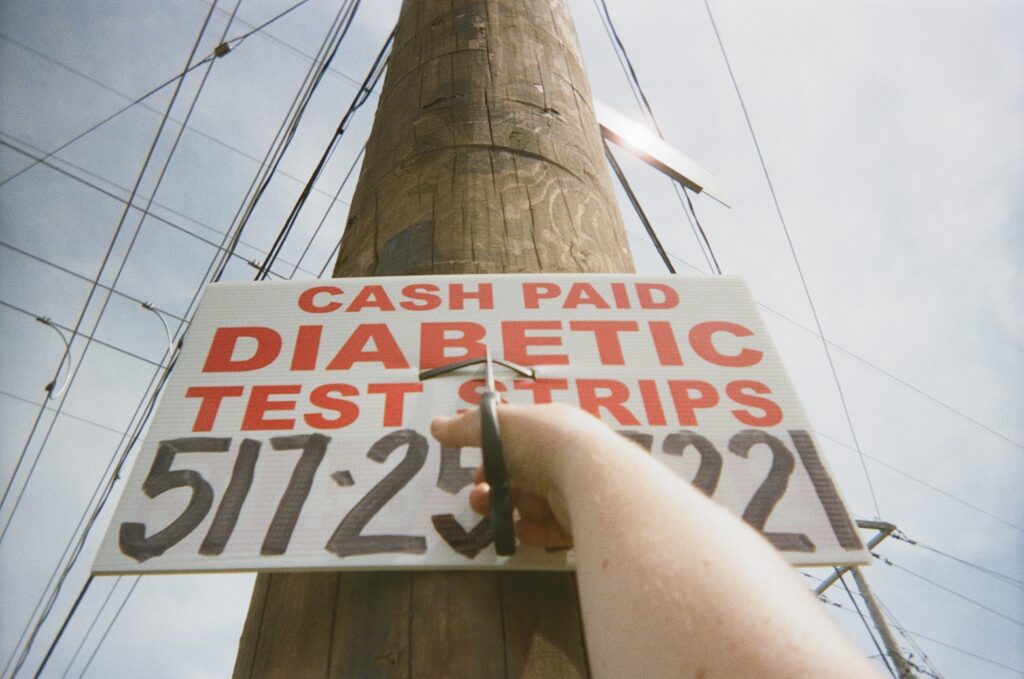 color photo of a hand with a scissors cutting down a red and black on white "cash paid diabetic test strips" sign that has been posted onto a roadside telephone pole. The photo is take from the perspective of the person doing the cutting.