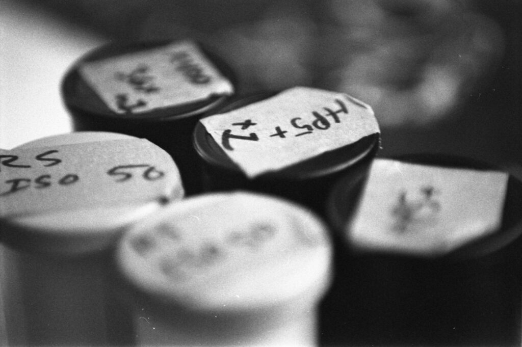 black and white image of 5 plastic film canisters with handwritten labels of the types of film and film speeds