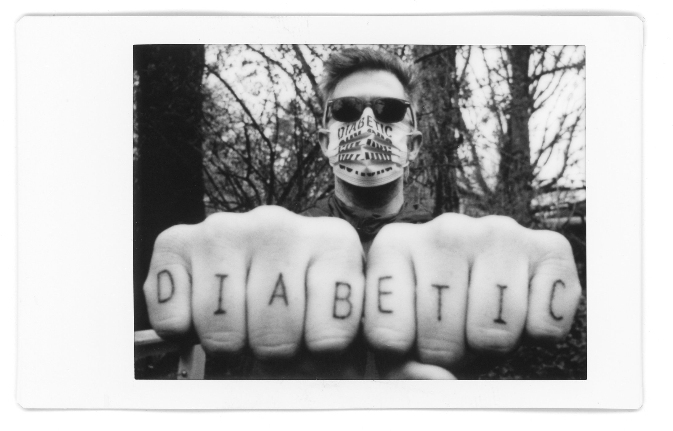 black and white instax photo of a white man wearing sunglasses and a surgical mask holding his fists out to the camera, showing that his knuckles are tattooed with "DIABETIC"