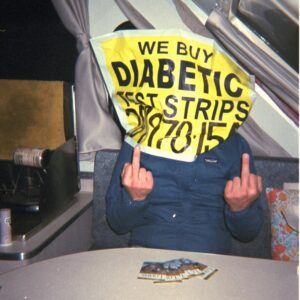 color photo of a person sitting at a table inside of a camper wearing a blue windbreaker jacket and a bag-like diabetic test strip sign over their head. both middle fingers are extended toward the camera.