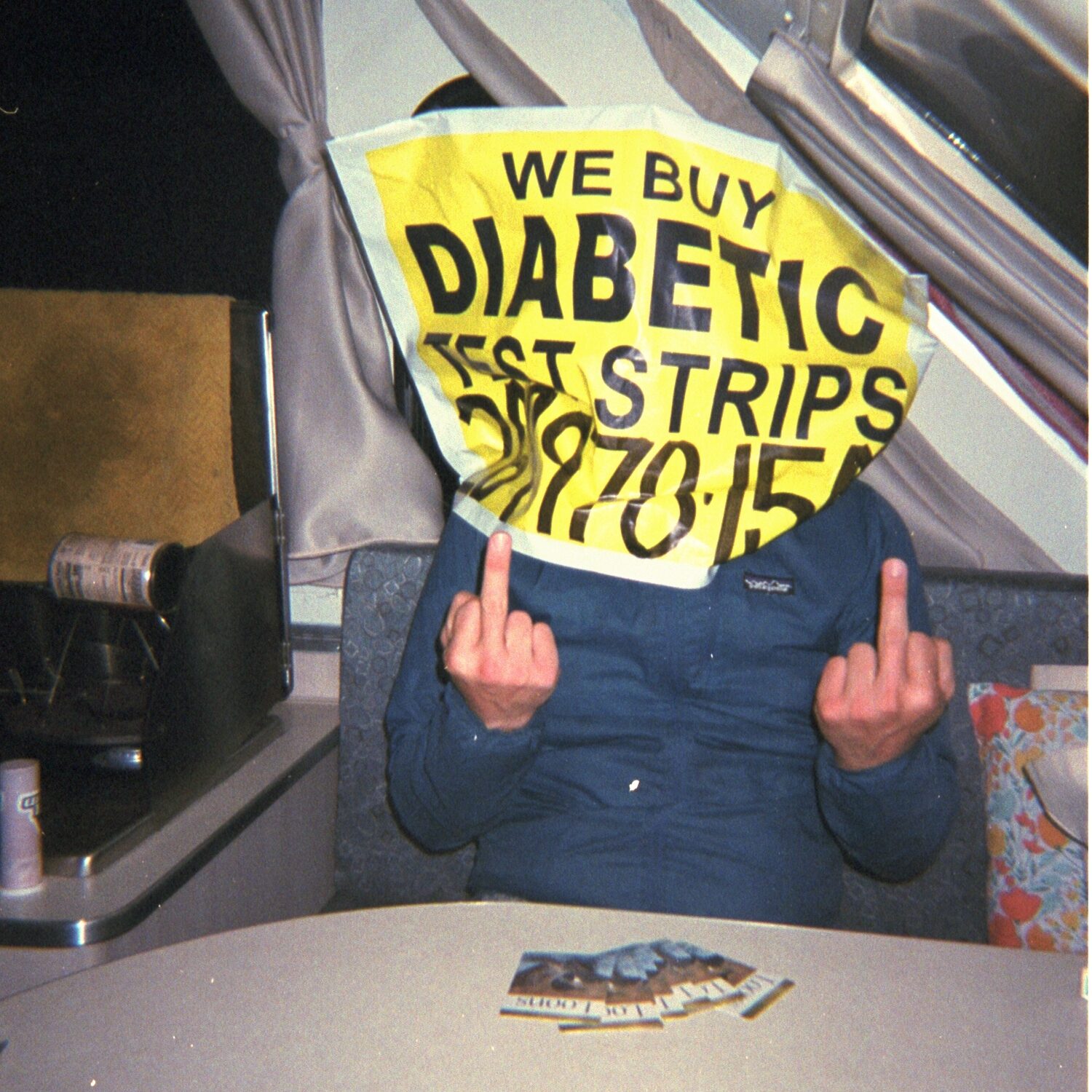 color photo of a person sitting at a table inside of a camper wearing a blue windbreaker jacket and a bag-like diabetic test strip sign over their head. both middle fingers are extended toward the camera.
